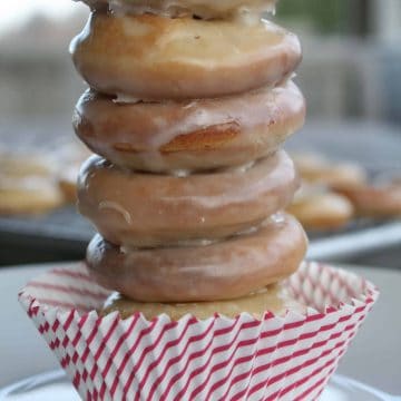 A stack of five glazed donuts balanced on top of a red and white striped cupcake liner, placed on a white plate, sits beside a gluten free single serve chocolate chip cookie for the perfect sweet pairing.