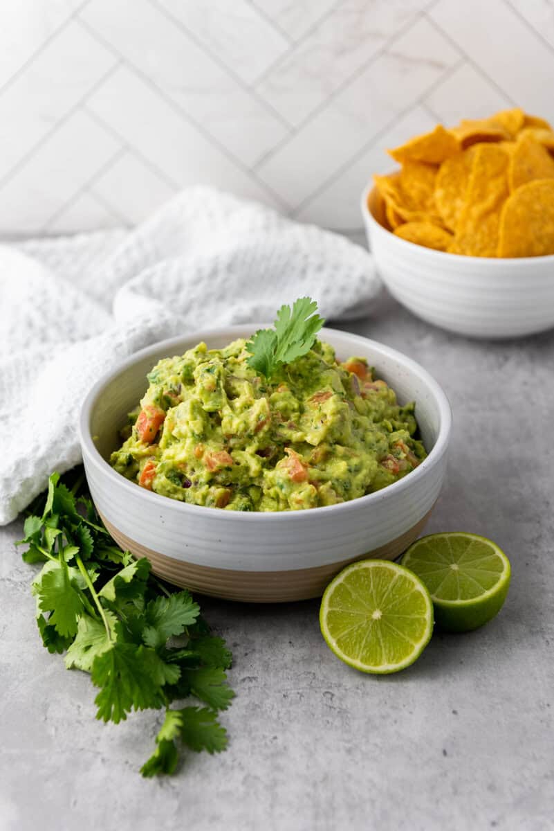 A bowl of addictive guacamole garnished with cilantro sits invitingly next to lime halves and a bowl of tortilla chips on a gray surface.