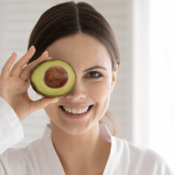 A woman holding an avocado in front of her eye, showcasing one of the 10 deliciously healthy foods that women can't get enough of.