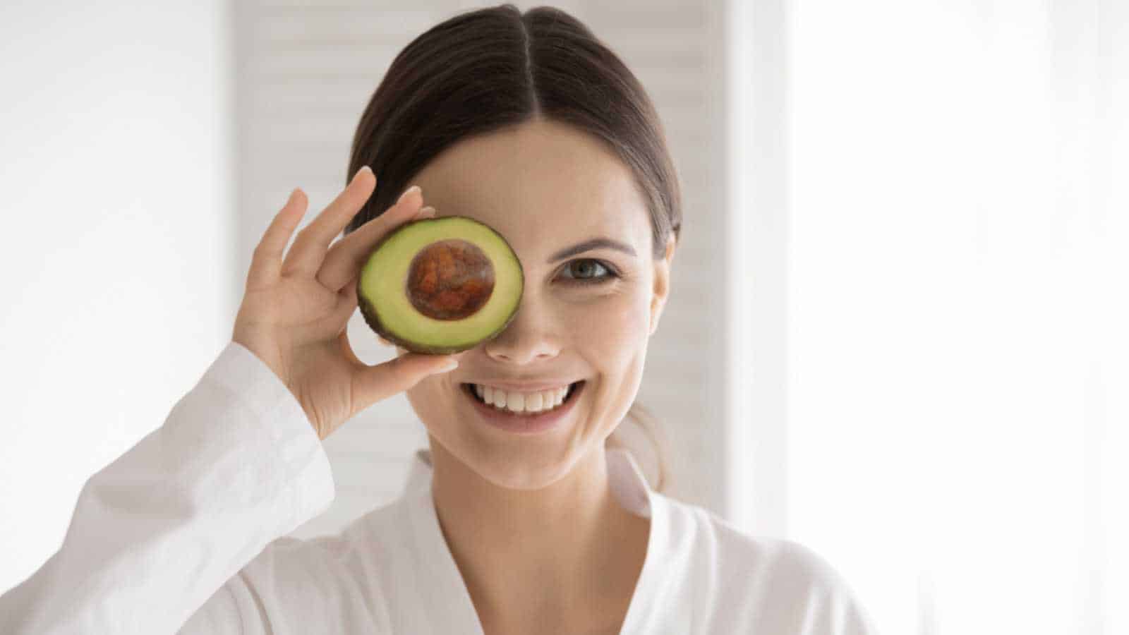 A woman holding an avocado in front of her eye, showcasing one of the 10 deliciously healthy foods that women can't get enough of.