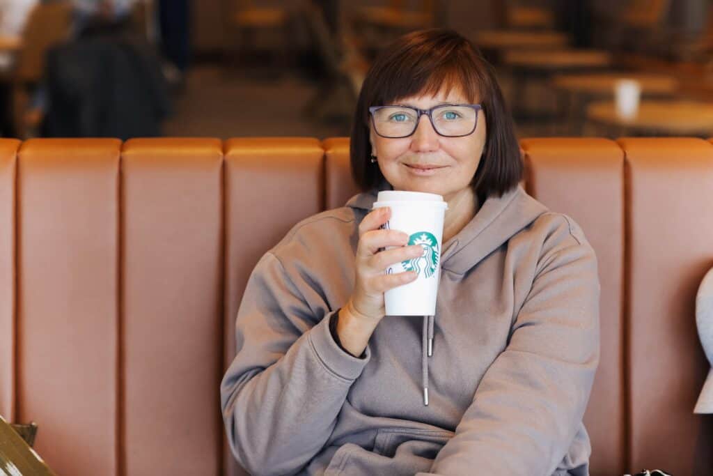 Smiling middle aged woman in casual clothes holds a paper cup of hot Starbucks coffee with straw at Starbucks coffee store at airport and waits for plane boarding.