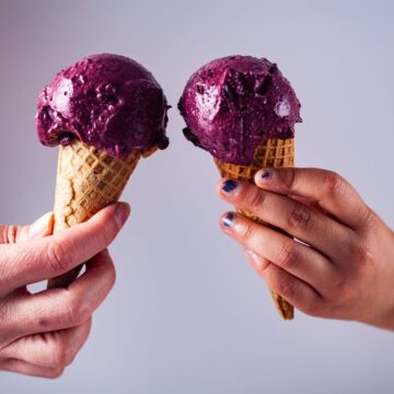 Two hands holding gluten-free ice cream cones filled with purple ice cream.