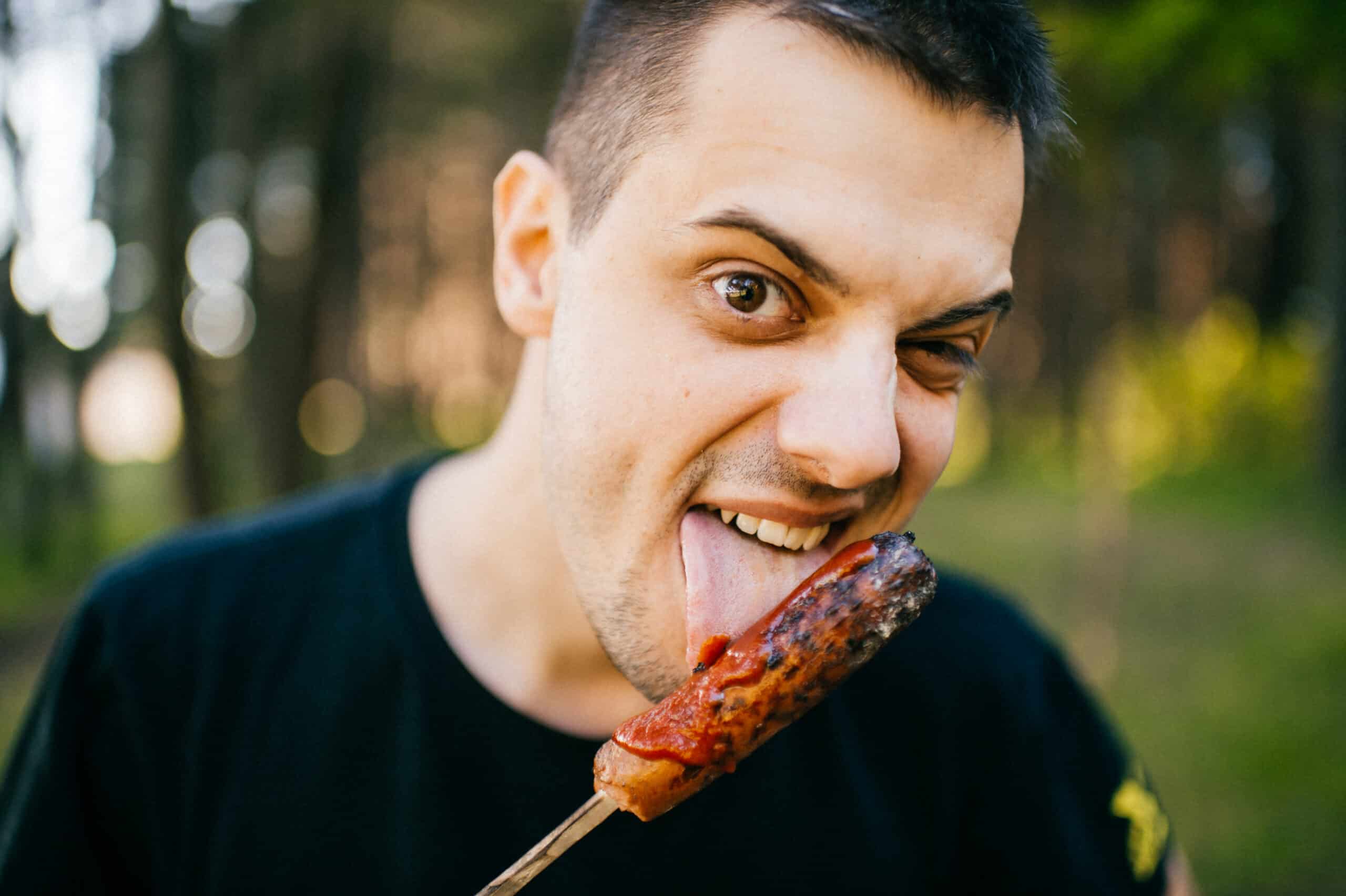 Closeup portrait of male licking and biting roasted sausage with disgusting ugly monstrous facial expression. Meat lover. Sinister evil diabolical face. Bum with maniacal eyes. Depth of filed. Funny