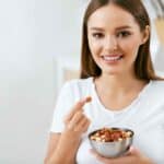 Healthy Food. Happy Woman Eating Nuts Holding Plate In Hands. Portrait Of Beautiful Smiling Female ON Diet With Raw Organic Almonds In Hands In Kitchen. Healthy Fats. High Quality