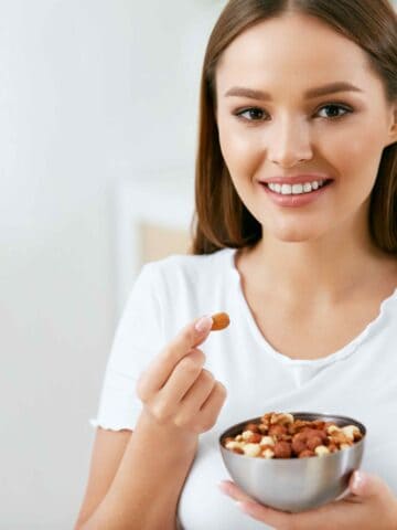 Healthy Food. Happy Woman Eating Nuts Holding Plate In Hands. Portrait Of Beautiful Smiling Female ON Diet With Raw Organic Almonds In Hands In Kitchen. Healthy Fats. High Quality