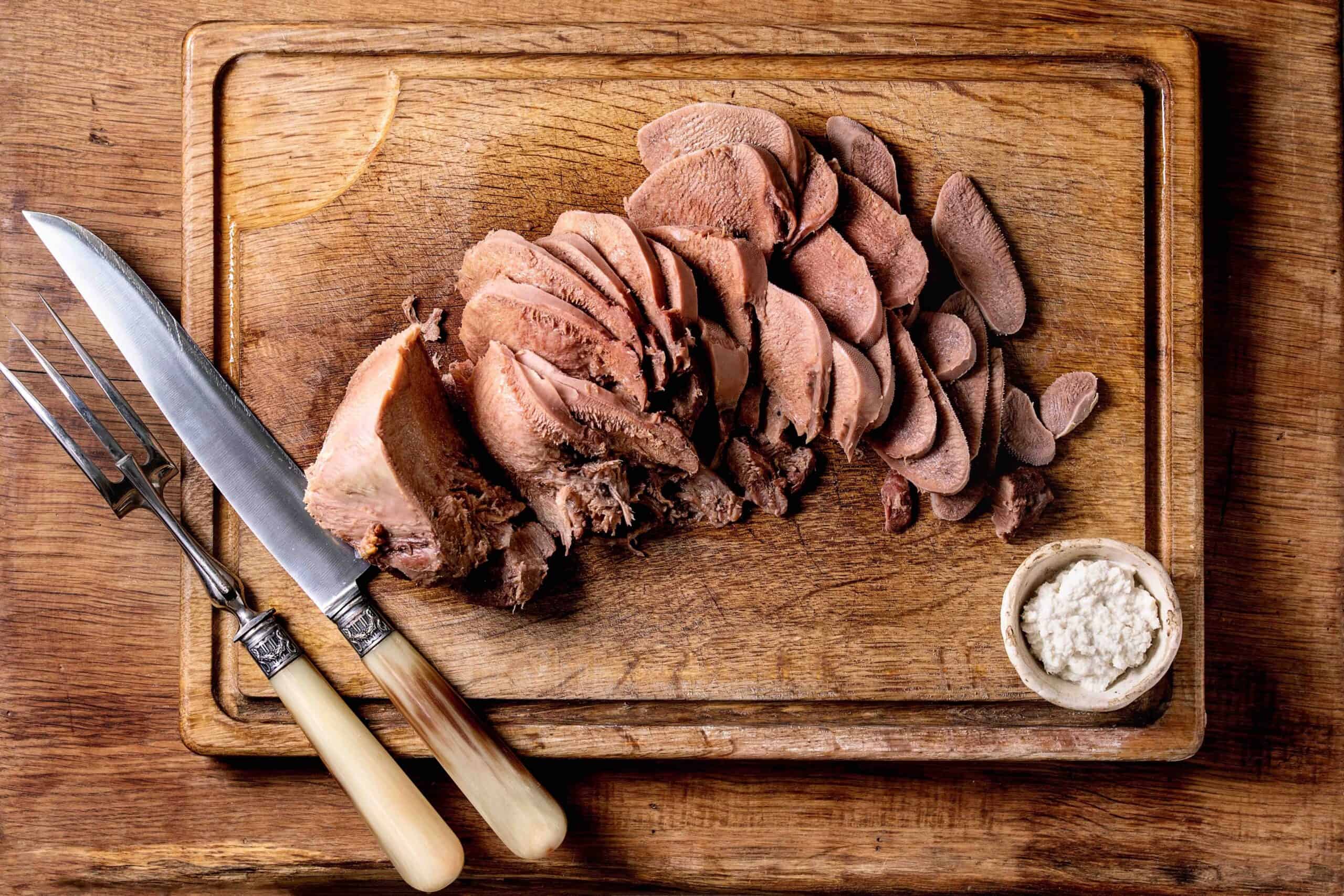 Baked beef tongue sliced, serving with horseradish sauce and meat knife on wooden cutting board over wood background. Flat lay.