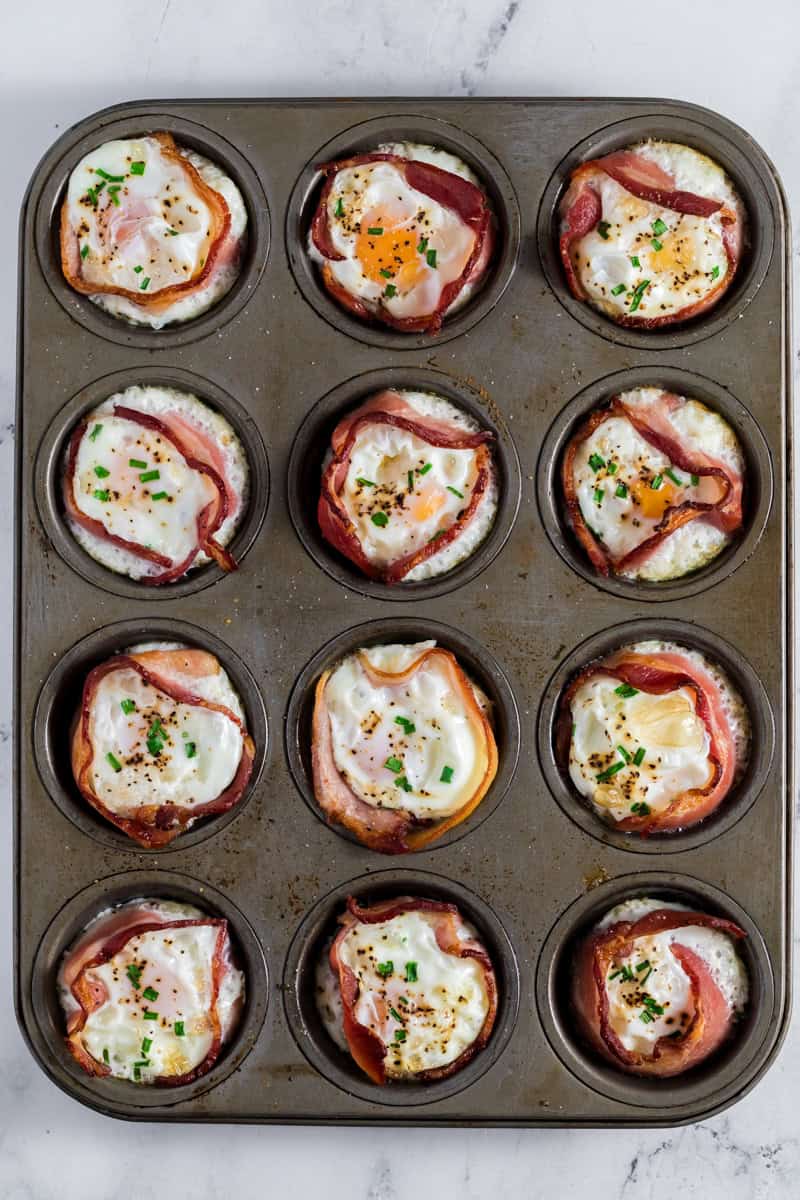 Muffin pan with twelve make-ahead bacon and egg cups, sprinkled with herbs, on a marble countertop.