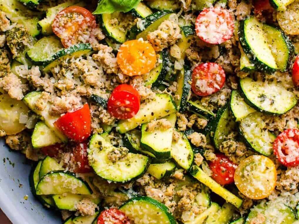 Close-up of a mixed vegetable salad featuring sliced zucchini, cherry tomatoes, and ground meat, garnished with grated cheese and herbs.