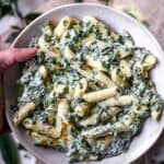 A hand holding a bowl of gluten-free pasta with spinach and cream sauce.