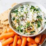 A bowl of gluten-free spinach dip with crackers and carrots.
