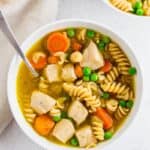 Gluten-free chicken noodle soup in a white bowl with carrots and peas.