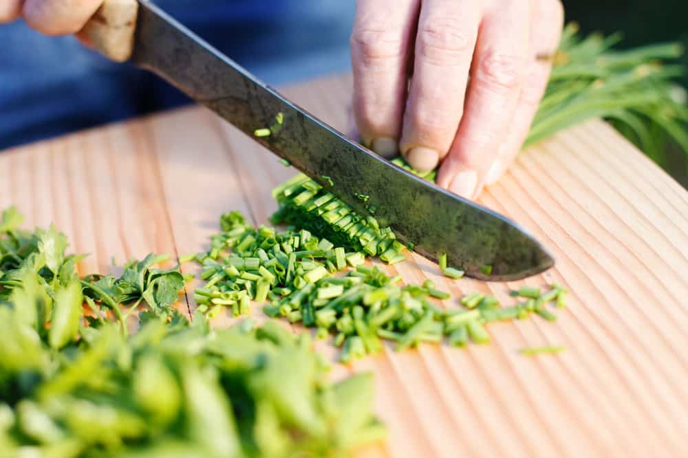Human hands cutting fresh chives using a knife and wooden chopping board