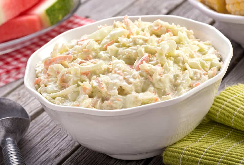 A bowl of delicious creamy homemade coleslaw on a rustic picnic table with watermelon and corn.