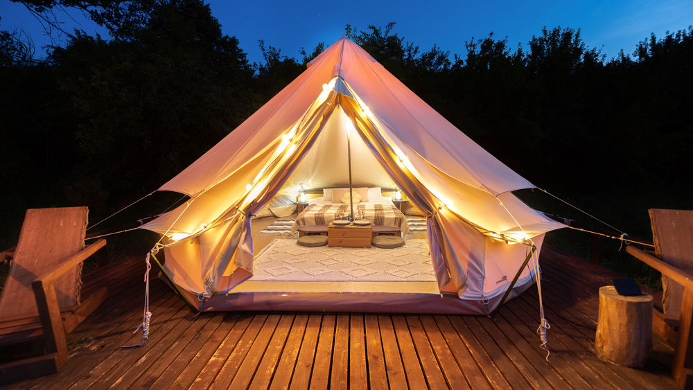 Tent interior and exterior with lamps and wooden chairs at glamping, forest around, night