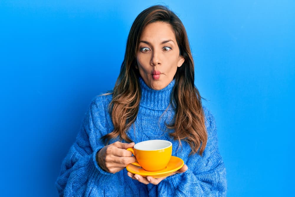 Young latin woman drinking a cup of coffee making fish face with mouth and squinting eyes, crazy and comical.