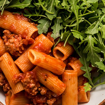 An Italian dinner recipe consisting of a bowl of pasta with flavorful meat sauce and topped with nutritious greens.