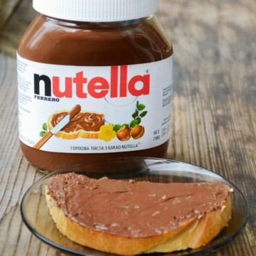 A jar of nutella sits next to a slice of bread.