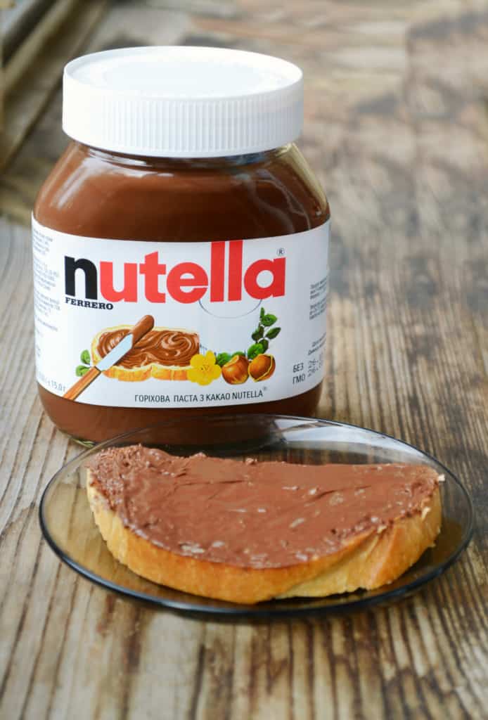 A jar of nutella sits next to a slice of bread.