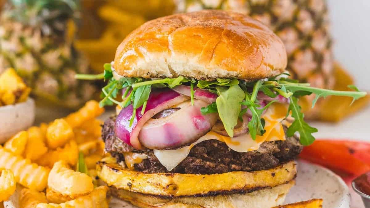 A burger with pineapple and onions served on a plate alongside fries.