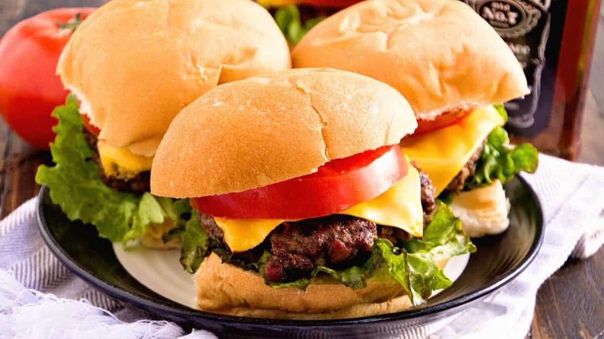 Three hamburgers on a plate alongside a bottle of bourbon.