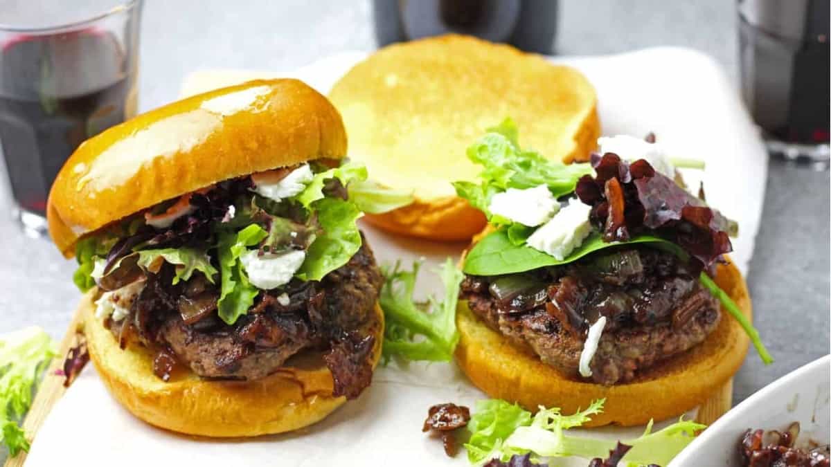 Two burgers on a plate next to a glass of wine and side dishes of Ham.