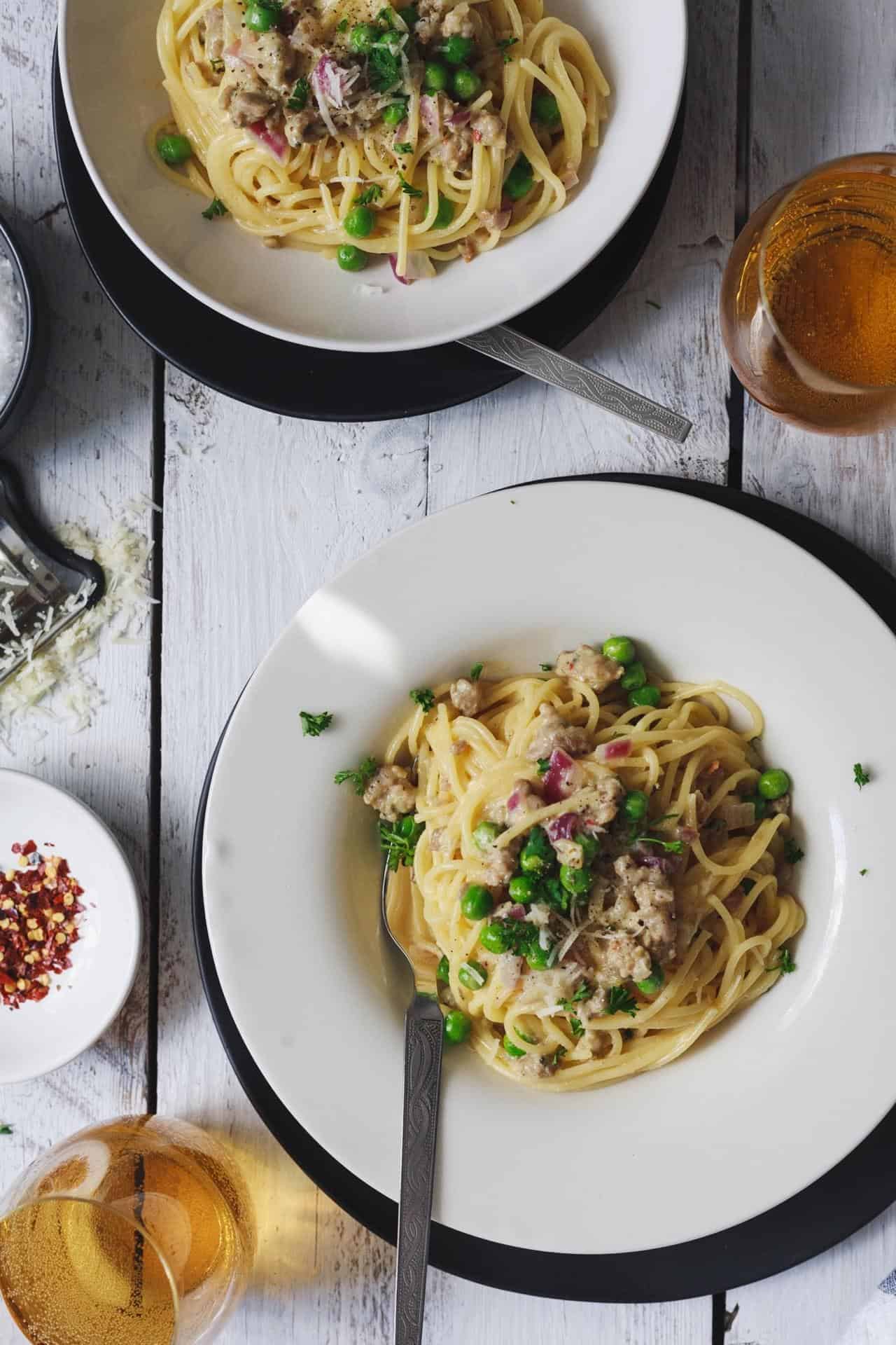 Two plates of spaghetti with sausage, peas, and meat on a wooden table.