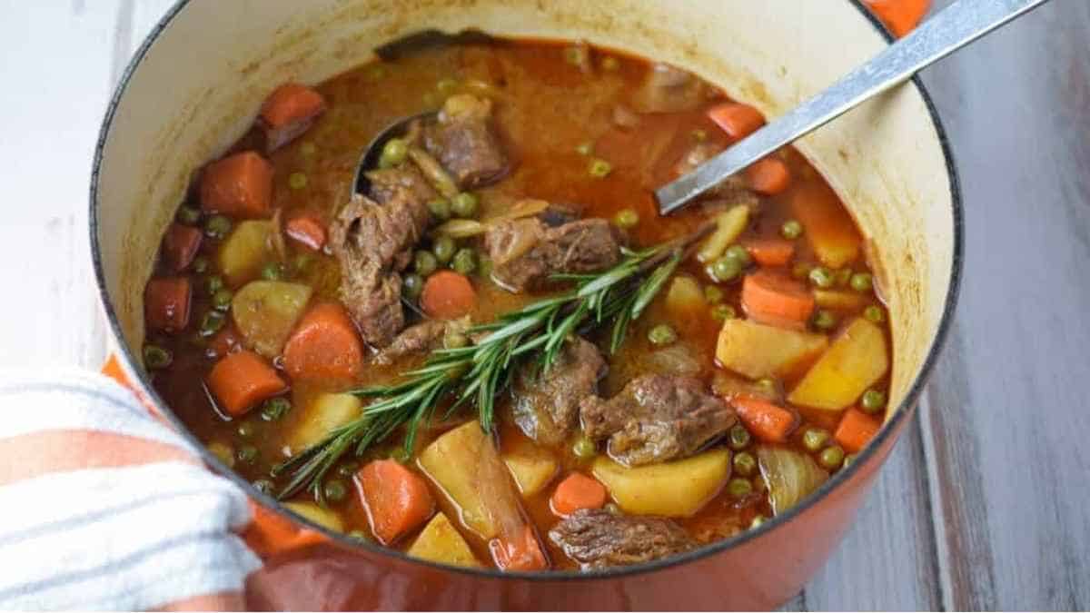 A pot of beef stew with vegetables and a sprig of rosemary.