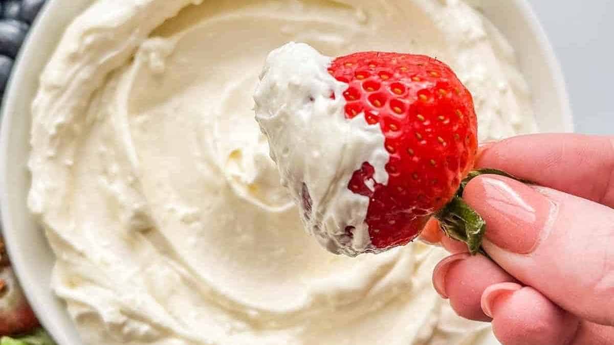 A hand is dipping a strawberry into a bowl of whipped cream.