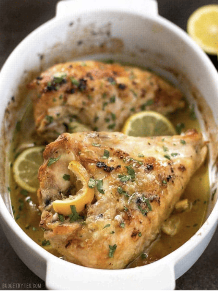 Bone-in chicken breasts in a white dish with lemons and herbs.