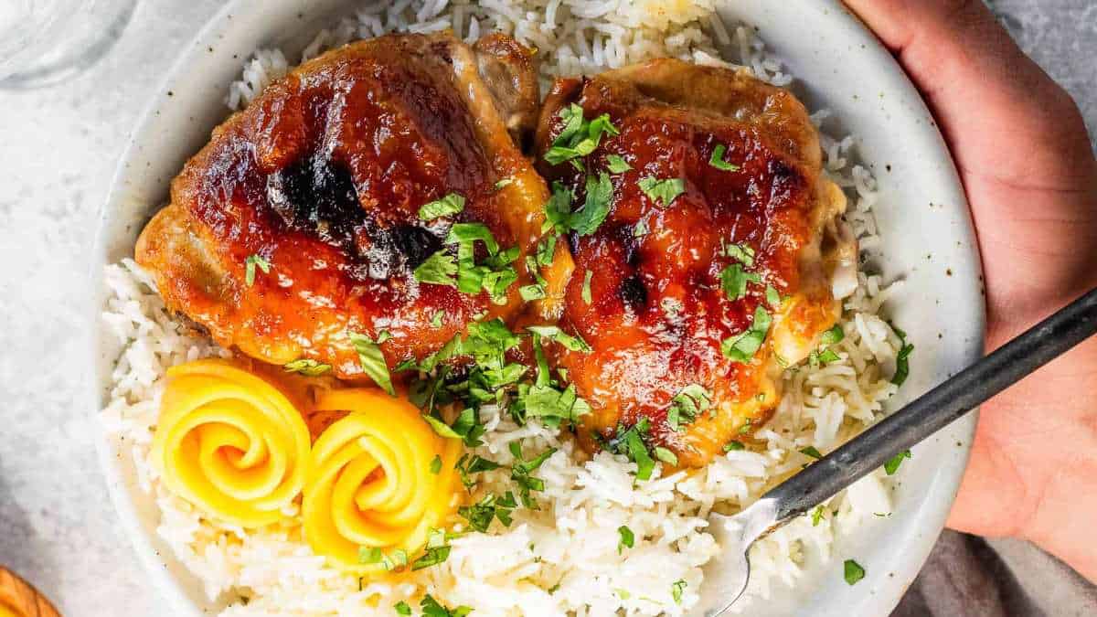 A hand holding a bowl of rice with chicken and mangoes on it.