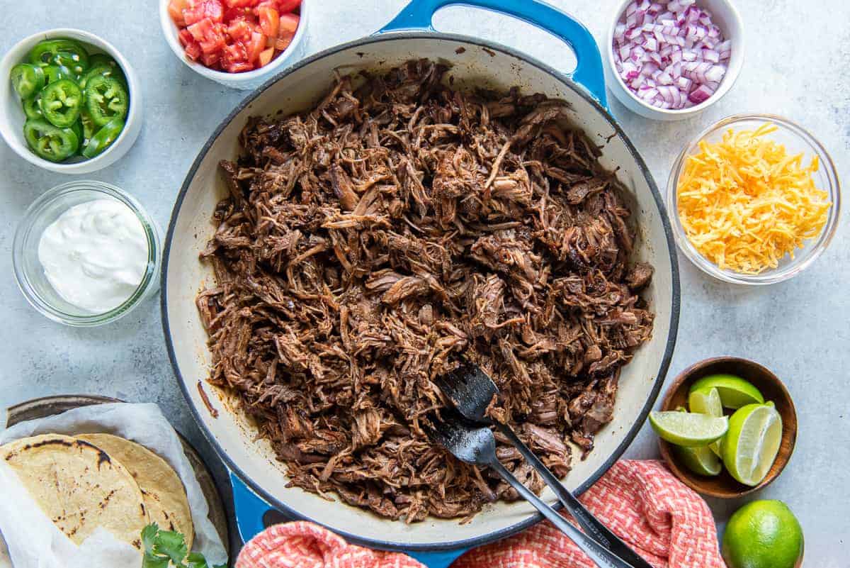 Mexican pulled pork in a skillet with sour cream, salsa, and tortillas.