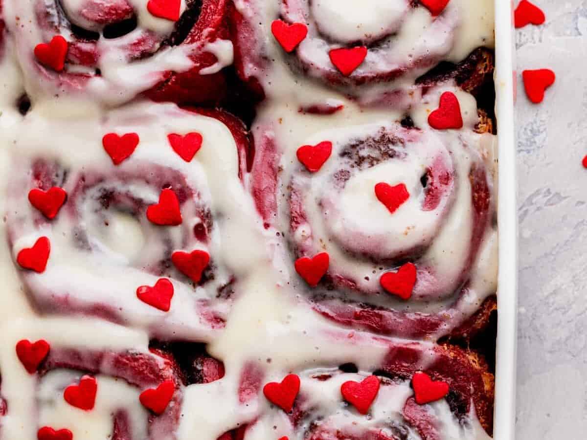 A tray of red velvet cinnamon rolls with white frosting and small hearts.
