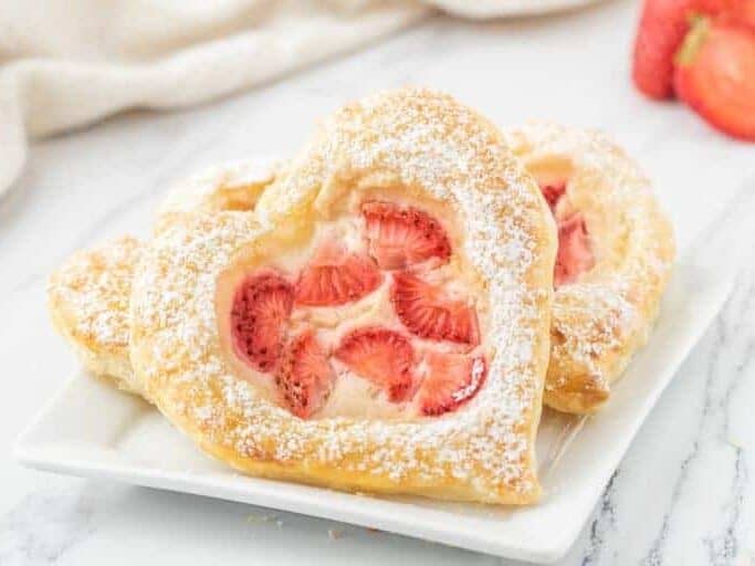 Two heart shaped pastries with strawberries and powdered sugar on a white plate.