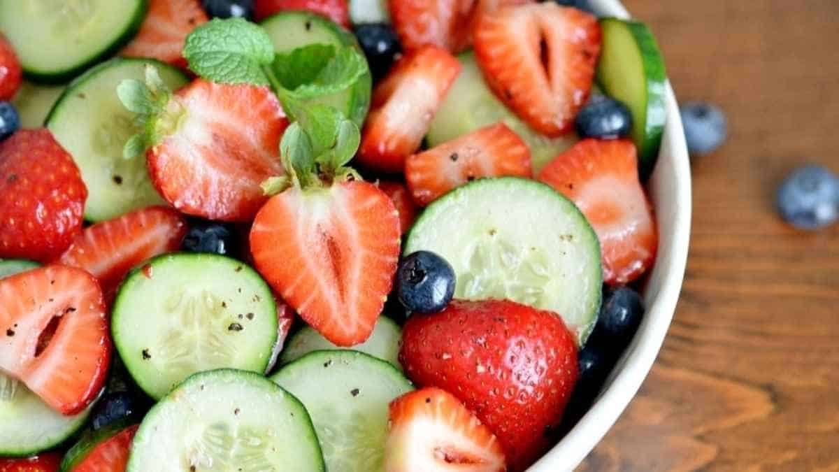 A bowl filled with cucumbers, strawberries and blueberries.