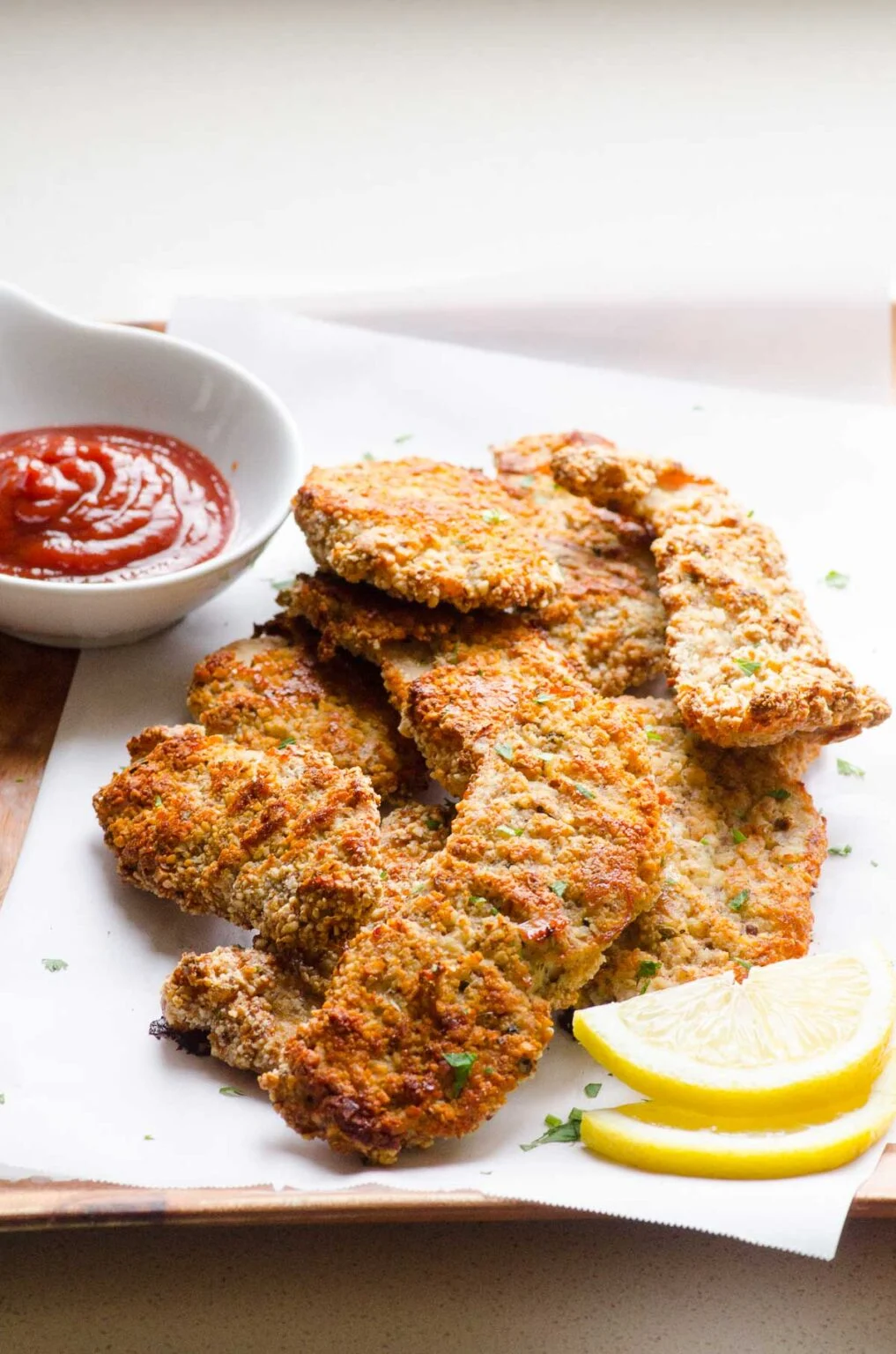 Chicken fried steaks with ketchup and lemon wedges on a wooden cutting board, perfect for chicken tender recipes.