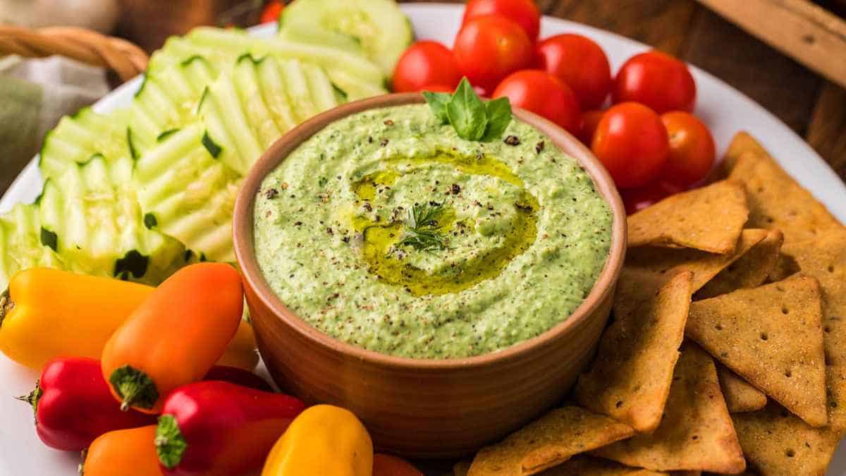 A bowl of green dip with crackers and vegetables.