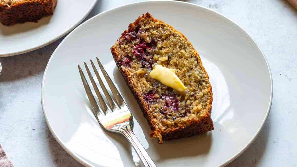 A slice of banana bread with berries and butter on a plate.
