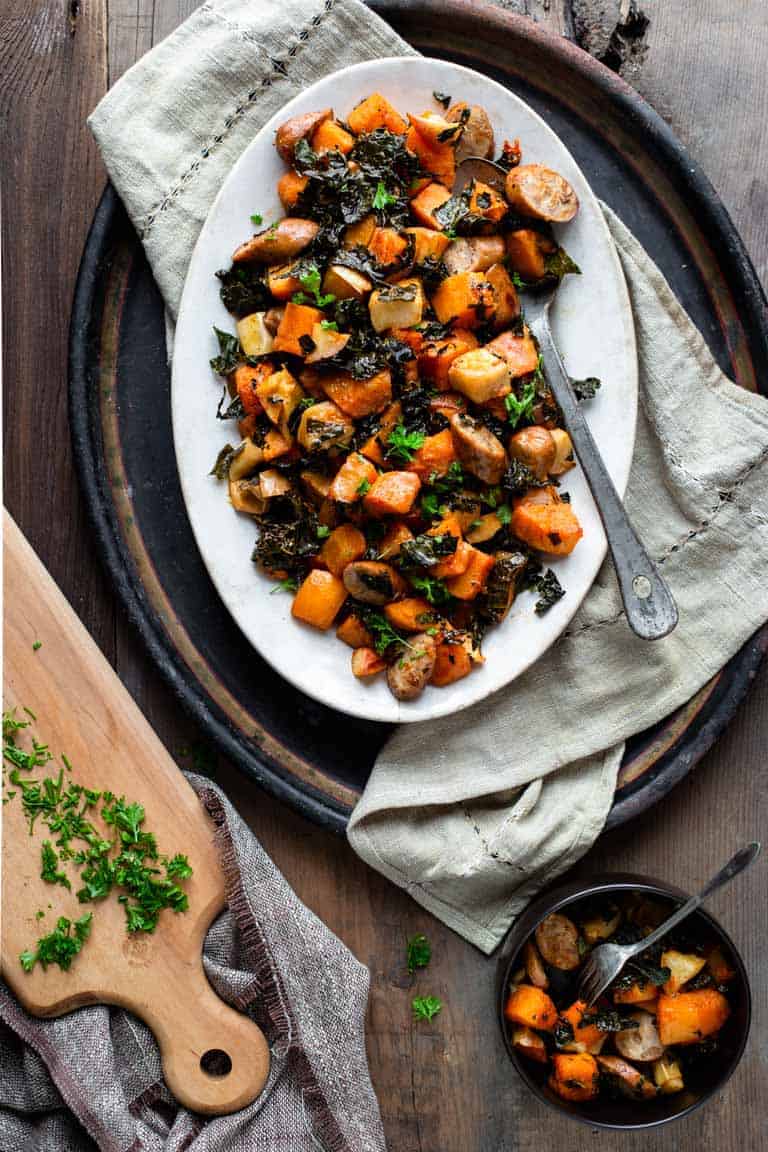 A plate of roasted sweet potatoes and kale topped with sausages on a wooden table.