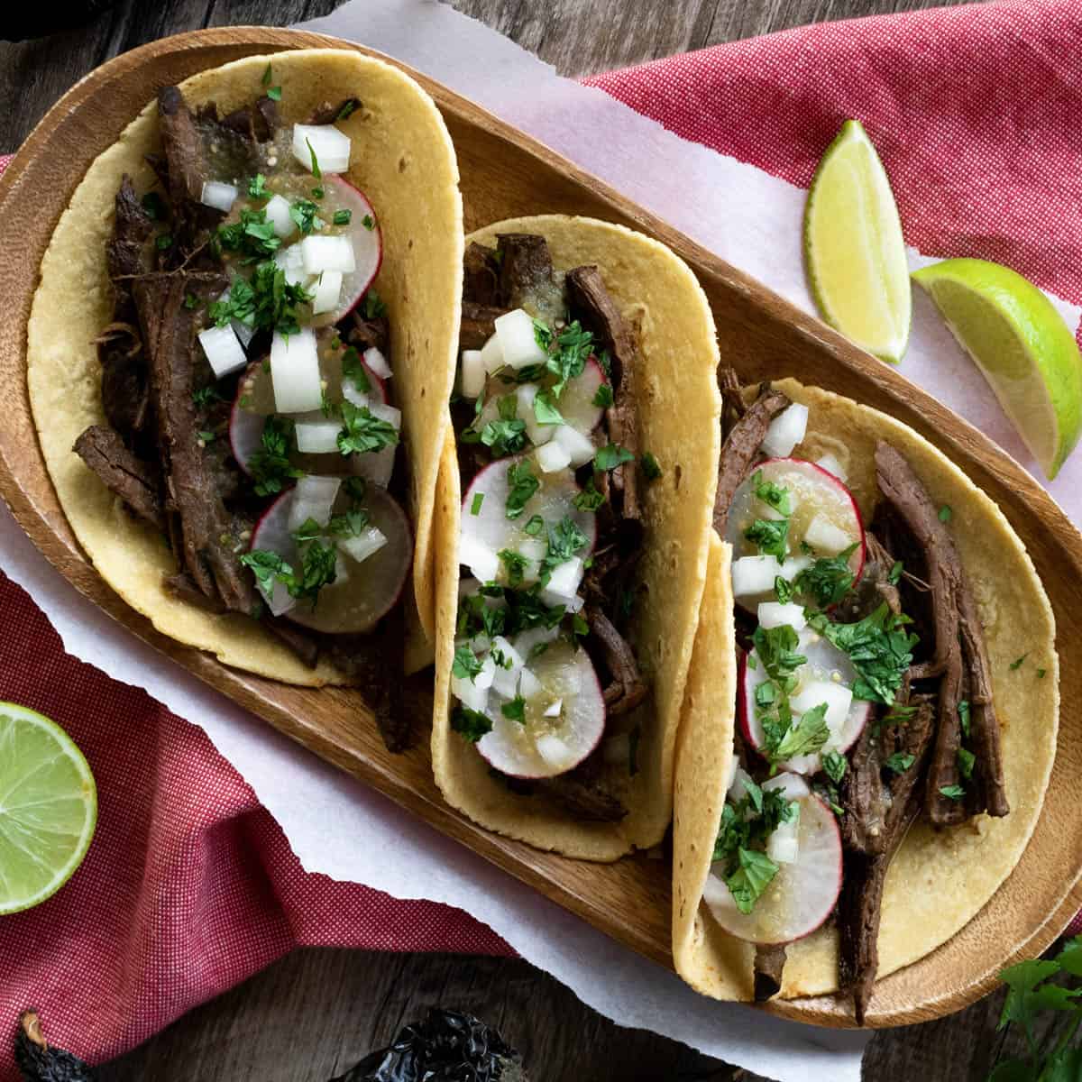 3 venison barbacoa tacos on wood plate.