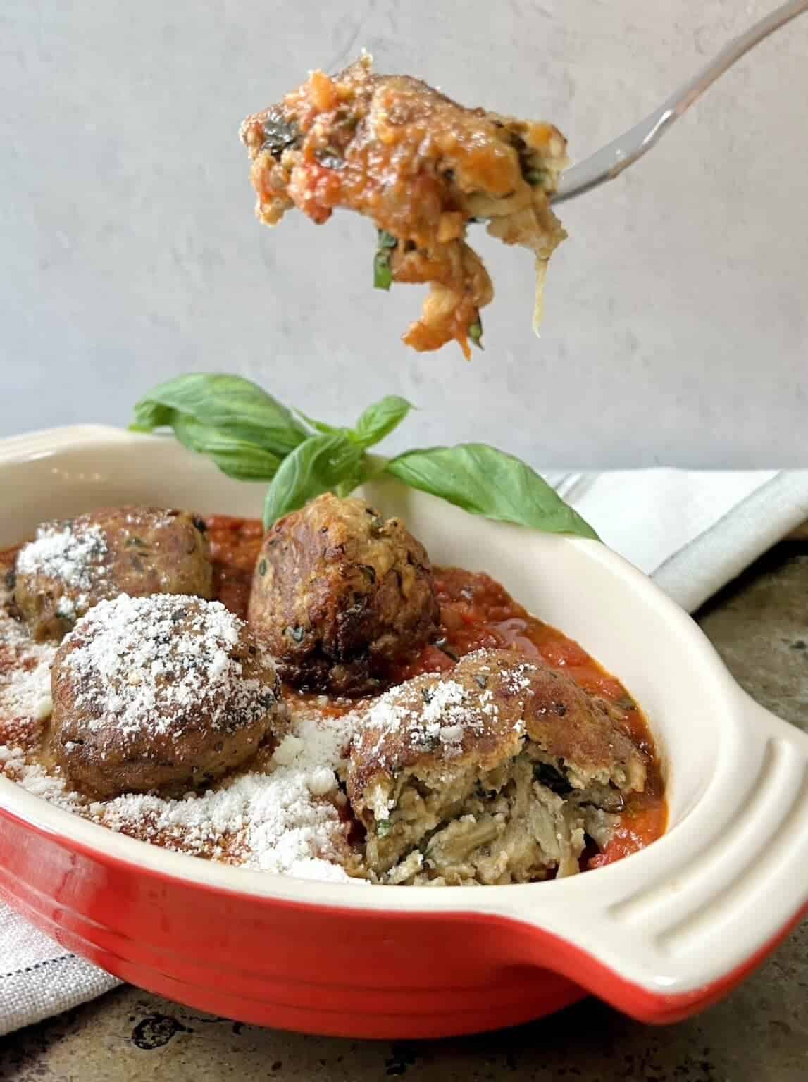 Forkful of polpette di melanzane over oval dish with basil garnish and grated cheese. 
