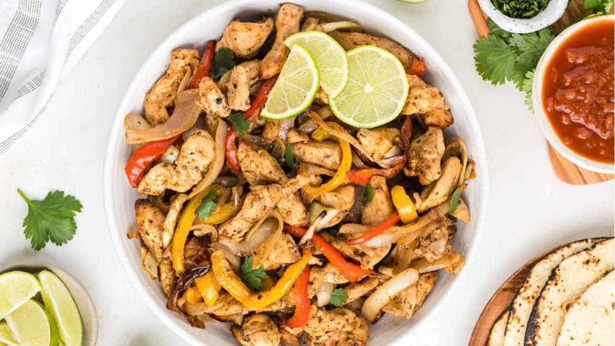 Chicken fajitas in a white bowl with limes and tortillas.