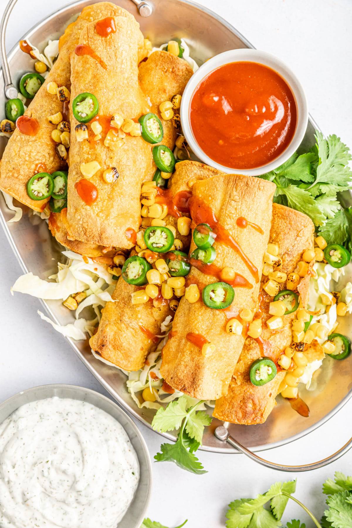 Overhead view of BBQ jackfruit taquitos piled onto a silver platter, topped with fire roasted corn, jalapeno slices, cilantro, and a drizzle of BBQ sauce. Small bowls of BBQ sauce and ranch dressing on the side.  