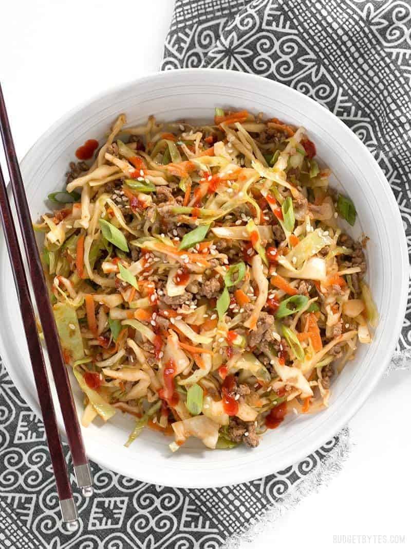 A bowl of Beef and Cabbage Stir Fry topped with sriracha, chopsticks on the side.

