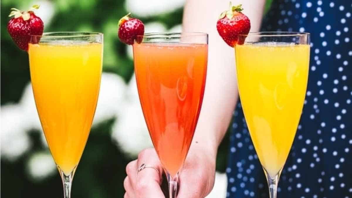 A woman is holding three glasses of orange juice and strawberries.