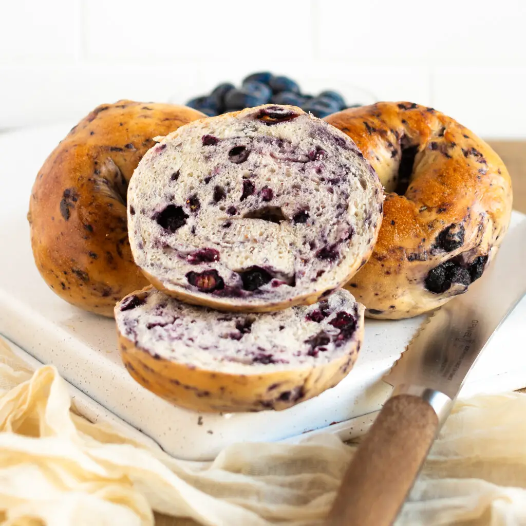Half a blueberry bagel, showing the inside, with a couple whole blueberry bagels and a bowl of blueberries in the background.
