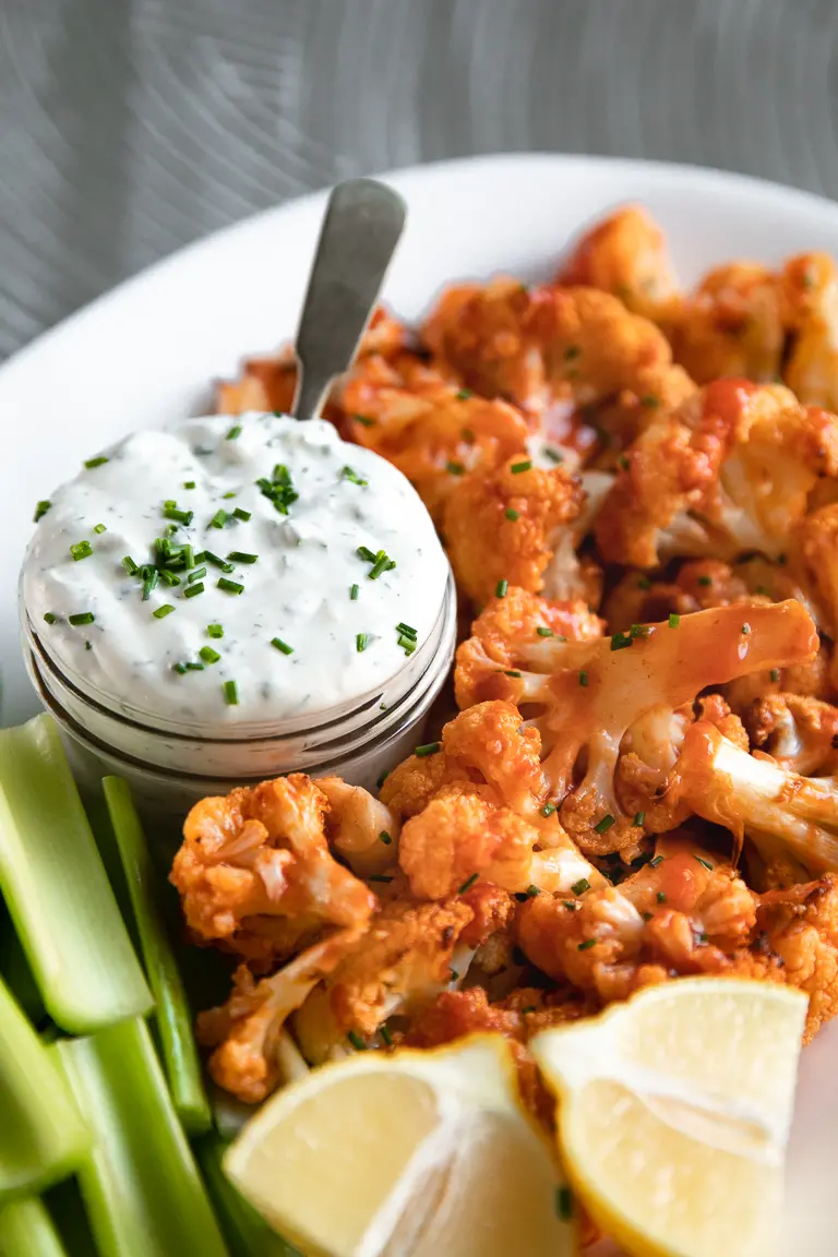Plate with baked buffalo cauliflower, jar filled with ranch dressing, celery sticks, and lemon wedges. 
