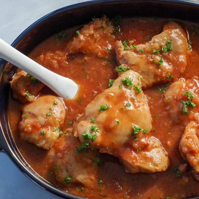 chicken in a hurry in a dark blue oval casserole dish, with a marble spatula, on a marble counter surface, with a grey oven mitt in the background.
