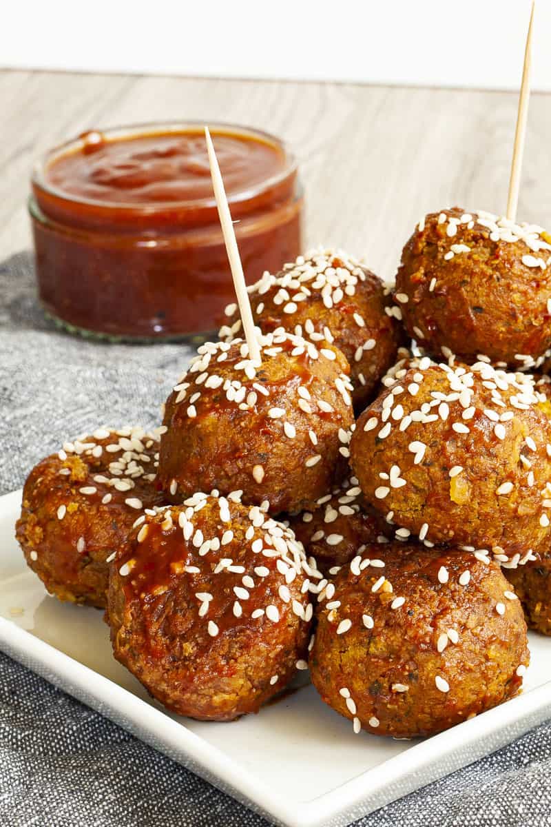 White plate with several brownish-redish balls on top of each other sprinkled with sesame seeds. In the background a small glass jar with red sauce.
