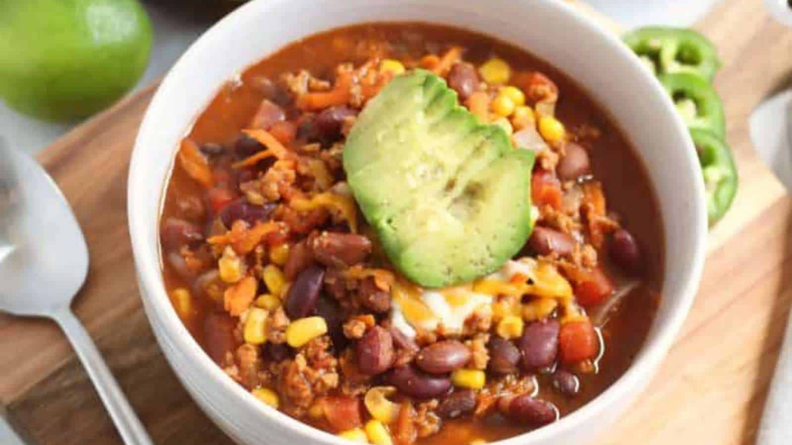 A bowl of chili with corn and avocado on a wooden cutting board.