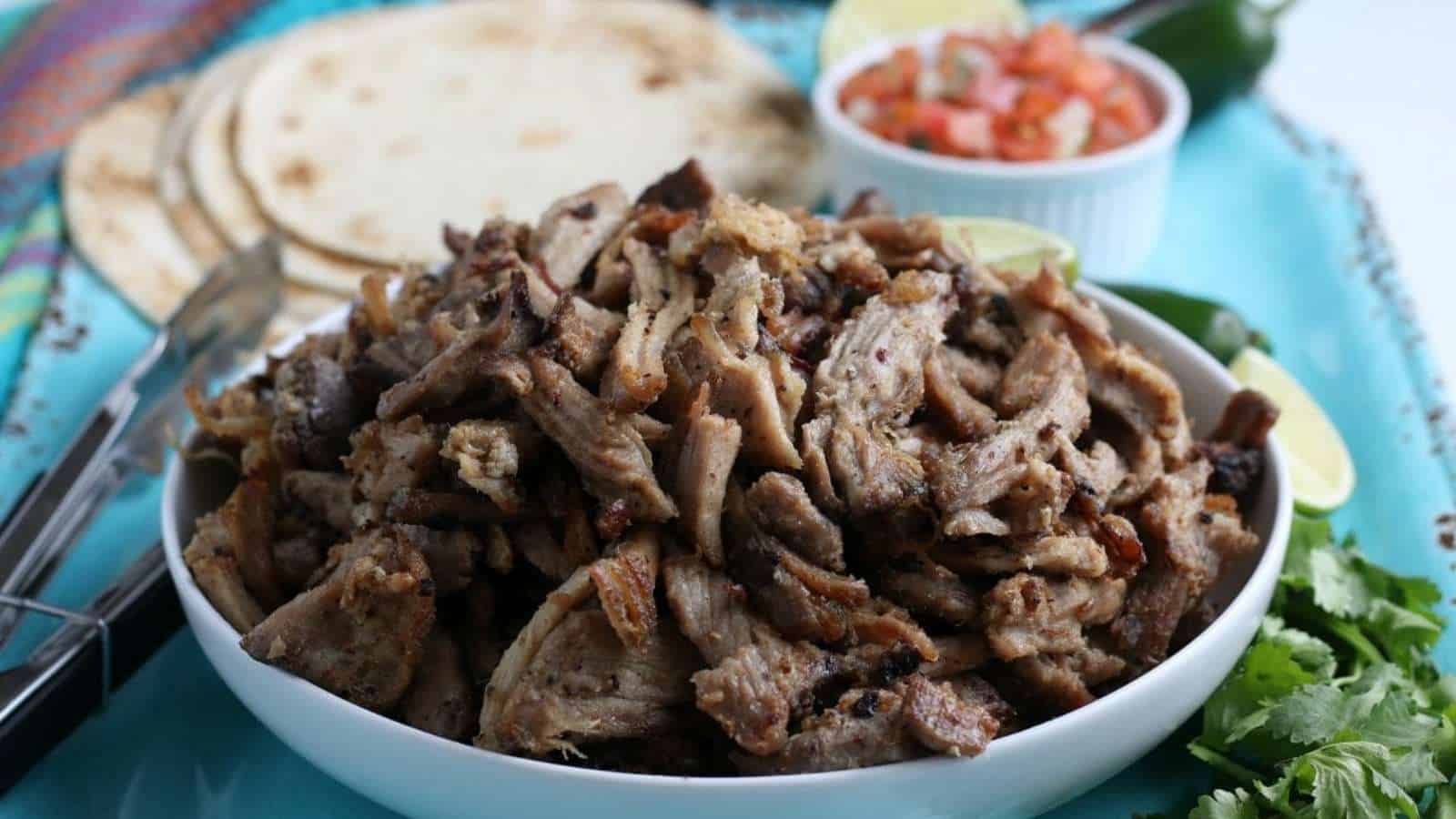 Shredded pork in a white bowl with limes and tortillas.
